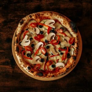 Top-down view of a freshly baked vegetarian pizza with mushrooms, tomatoes, olives, and red peppers on a wooden table.